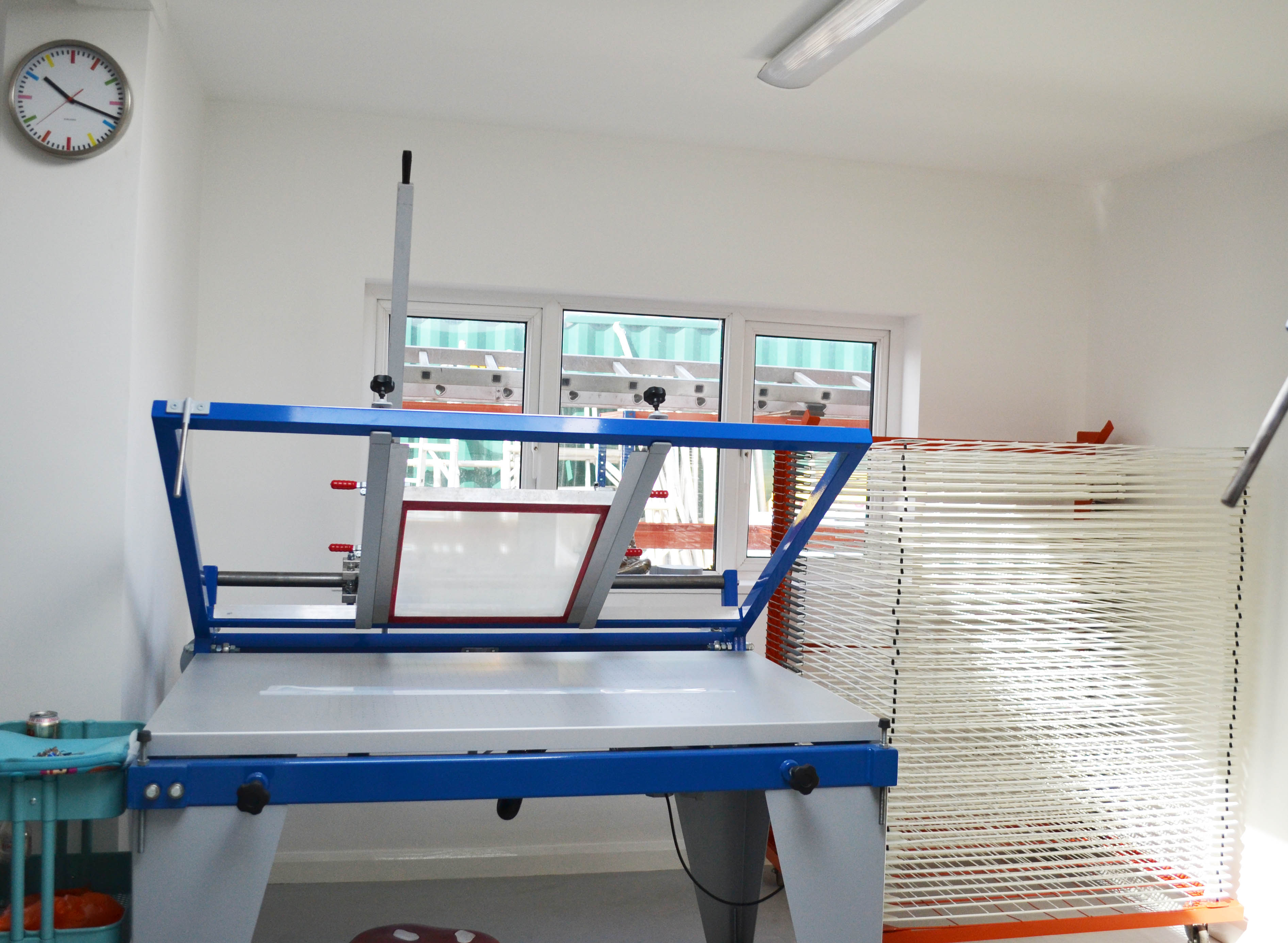 Dye Sublimation vs Screen Printing for Cosplay: Which One Wins? 3 A screen printing hand bench in a studio with a mesh screen frame mounted on hinge clamps and a drying rack beside it.